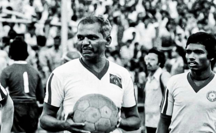 Close-up vintage image of Tulsidas Balaram in his football jersey, holding a soccer ball with a large crowd in the background.